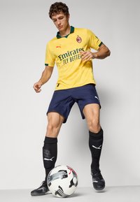 Gul fotbaltrøye med grønn krage og logo, marineblå shorts, svarte sokker og svarte fotballsko. Spilleren står med en mønstrete fotball.