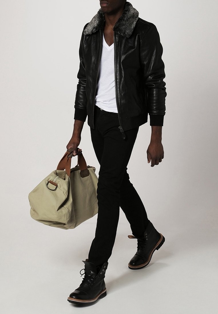 Homme portant une veste en cuir noire avec col en fourrure, t-shirt blanc à col en V, pantalon noir, bottes noires, portant un sac de voyage beige avec poignées marron.