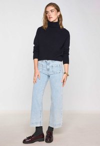 Donna che indossa un maglione a collo alto nero, jeans a gamba larga blu chiaro, calzini scuri e mocassini bordeaux, in piedi su uno sfondo uniforme.