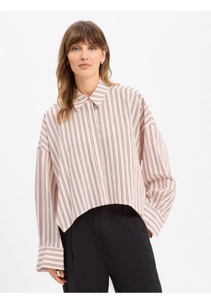 Femme portant une chemise à rayures rose clair à manches longues avec de larges poignets et un pantalon noir, debout devant un fond blanc uni.