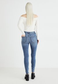 Kék skinny farmernadrág magas derékkal, hátsó zsebekkel és varrási részletekkel. Egy illeszkedő, fehér, vállat szabadon hagyó toppal és fekete magassarkú cipővel párosítva.