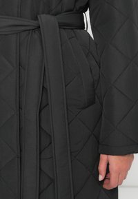 Tamsiai pilkas siuvinėtas chalatas su įstrižais siūlėmis, diržo juosta, lygiu paviršiumi ir ilgomis rankovėmis, kuriame pabrėžiamas rankos ir diržo artimas planas.