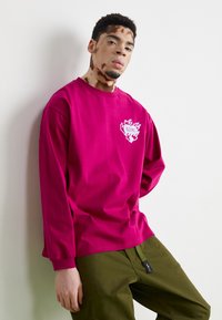 Fuksjowa koszulka z długim rękawem z białym wzorem na lewej piersi, w połączeniu z oliwkowymi spodniami z metką logo.