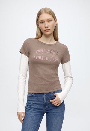 Jeune femme aux longs cheveux blonds portant un haut marron superposé à manches courtes et un haut blanc à manches longues avec un jean bleu, debout devant un fond blanc.