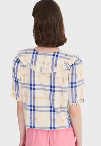 Femme portant un chemisier crème à carreaux bleus et oranges, avec un col et des manches à volants, assorti à un short rose, vue de dos.
