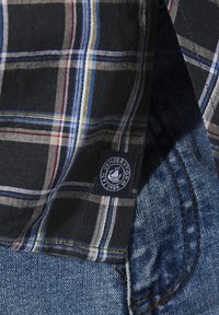 Schwarz-karierter Hemd mit weißen, roten und blauen Streifen, mit einem Stoffetikett, das das Branding zeigt; kombiniert mit blauen Jeans.