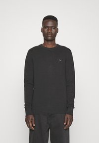 Pullover nero a maniche lunghe con un design a coste testurizzato e scollo rotondo. Presenta un piccolo logo sul petto e polsini aderenti.