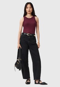 Mujer con una blusa sin mangas color burdeos, pantalones negros de tiro alto con cinturón, zapatillas negras puntiagudas, sosteniendo un bolso negro por la correa.