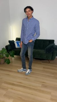 Homme portant une chemise à carreaux bleue, un jean déchiré et des baskets, debout avec une main dans la poche dans un salon avec un canapé vert et des plantes.