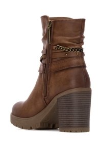 Stivale alla caviglia marrone in ecopelle con tacco spesso e scanalato. Presenta una cerniera laterale e accenti decorativi a catena intorno alla parte superiore.