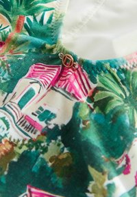Farverigt mønstret stof med tropiske motiver i grøn, pink og hvid, med en gulddetalje ved trykknappen nær halsudskæringssømmen.