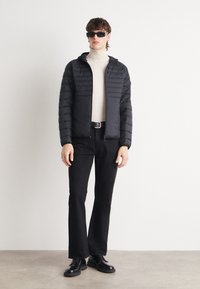 Svart puffjacka med horisontellt quiltat mönster, dragkedja framtill och huva, bärs över en beige polotröja, ihop med svarta utsvängda byxor.