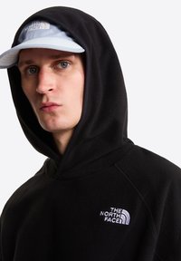 Μαύρο φλις φούτερ με μεγάλο κουκούλι και κεντημένο λευκό λογότυπο στο στήθος. Ανοιχτό μπλε καπέλο με επίπεδη γείσο φορεμένο πάνω από το φούτερ.