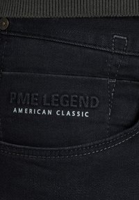 Los pantalones de mezclilla negros presentan una tela texturizada con costuras visibles. Una etiqueta muestra "PME LEGEND AMERICAN CLASSIC" en letras en relieve.