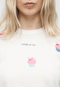 Ξανθιά γυναίκα που φοράει λευκό πουκάμισο με ροζ σχέδια κυπέλων κέικ και το κείμενο "CHERRY ON TOP" τυπωμένο πάνω από το κεντρικό κύπελλο.