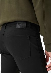 Pantaloni neri con una vita testurizzata e un'etichetta del marchio rettangolare. I pantaloni hanno due tasche posteriori con dettagli di cucitura visibili.