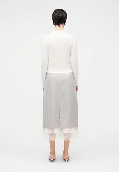 Fehér hosszú ujjú pulóver, rövidített fazonban, szürke bokáig érő szoknyával párosítva, amely varrással és alulról látható fehér csipkés díszítéssel rendelkezik.