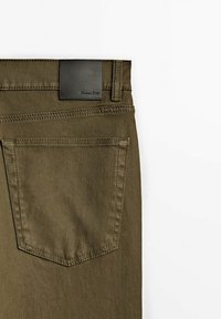 Olivgrüne Hose mit glatter Oberfläche, ausgestattet mit einem schwarzen Lederetikett am Bund und einer einzigen aufgesetzten Gesäßtasche. Sichtbare Ziernähte.