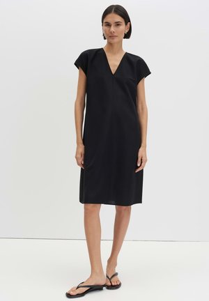 Woman with short dark hair wearing a simple black knee-length dress and black flip-flop sandals standing against a plain white background.