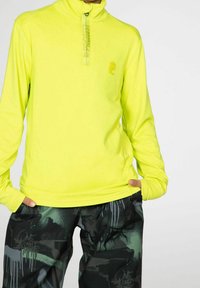 Suéter amarillo de manga larga con cuello alto, que presenta un cierre de cremallera y un detalle de logo. Combinado con pantalones oscuros estampados que muestran un diseño gráfico.