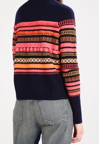 Pull bleu marine avec des rayures géométriques roses, oranges et jaunes, orné de pompons, porté avec un jean en denim bleu clair.