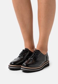 Chaussures en cuir verni noir à lacets, présentant un design à bout aile, des détails perforés et une semelle superposée contrastante avec une accentuation en brun clair.