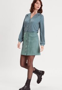Blouse bleue à motifs avec des manches larges, associée à une jupe en daim verte à boutons. Le modèle porte des bottines noires et des collants noirs transparents.