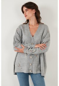 Cárdigan gris de punto con escote en V, detalles florales bordados y botones grandes. Presenta mangas anchas y un corte holgado.