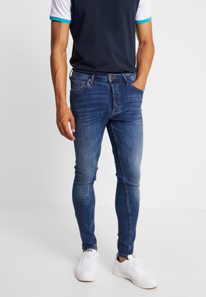 Mężczyzna w obcisłych niebieskich dżinsach z lekkim przetarciem i białych trampkach, stojący z jedną ręką w kieszeni na białym tle.