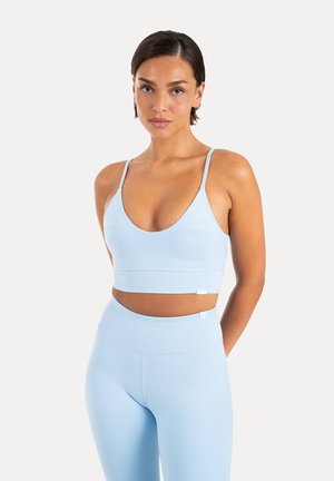 Kvinde med kort mørkt hår iført lyseblå sports-bh og matchende højtaljede leggings, stående mod en ensfarvet hvid baggrund.