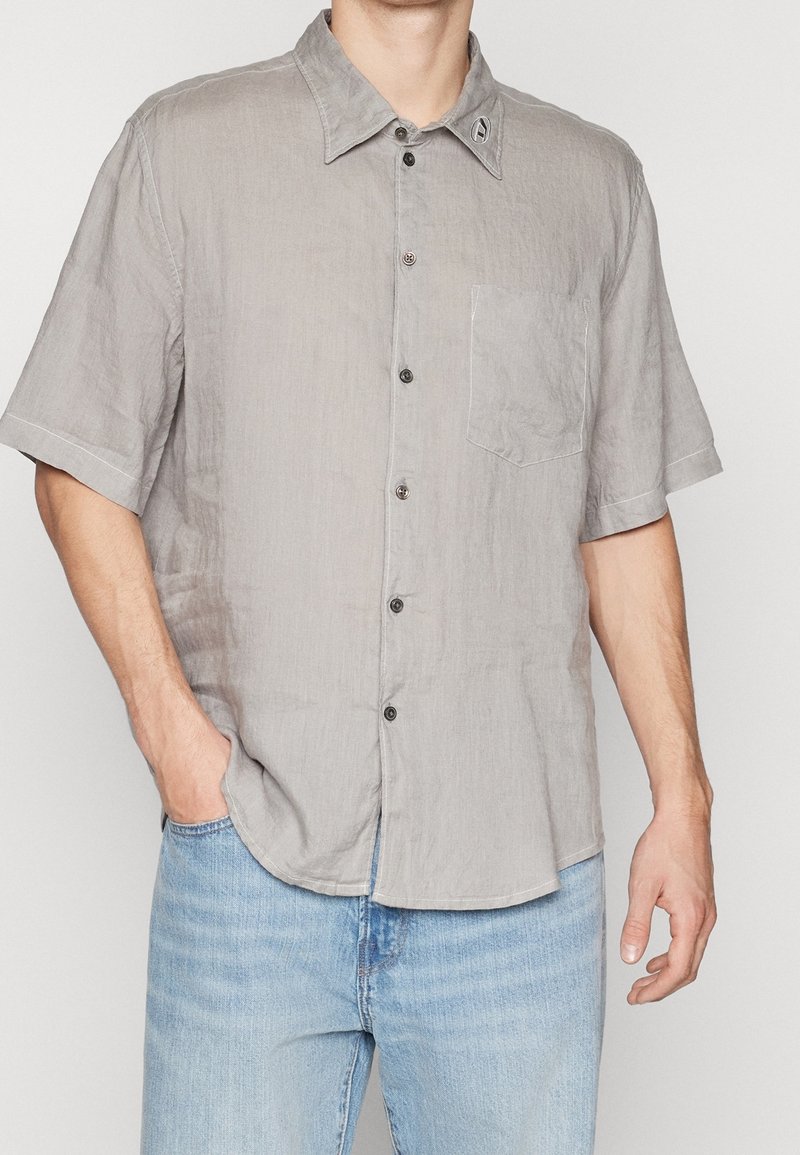 Homme portant une chemise à manches courtes gris clair avec une poche poitrine et un jean bleu clair, debout avec une main dans la poche.