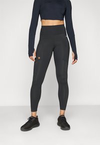 Zwarte leggings met hoge taille, twee zijzakken en een klein bruin logo. Gemaakt van rekbaar materiaal met een gladde textuur.