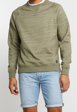 Mann trägt olivgrünes Langarm-Sweatshirt und hellblaue, hochgekrempelte Jeansshorts, steht vor einem schlichten Hintergrund.