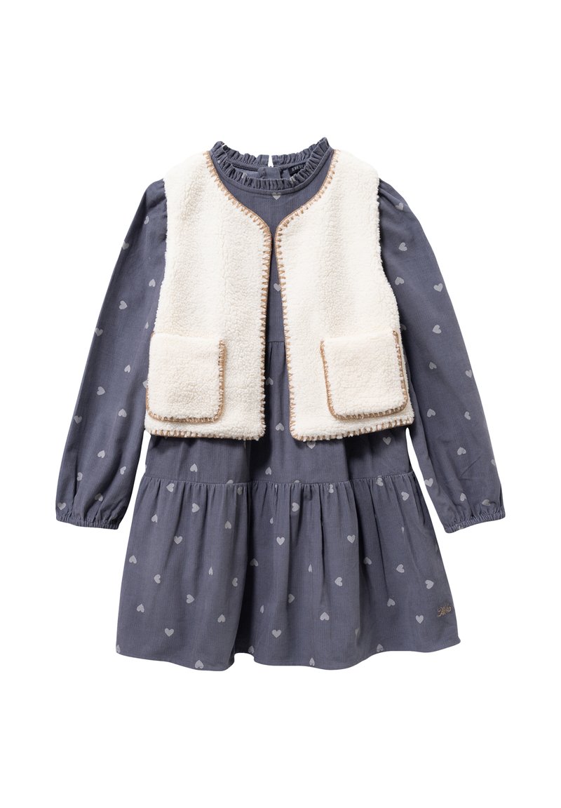 Vestido azul marino con estampado de corazones, mangas largas y falda fruncida, combinado con un chaleco de piel sintética color crema con ribetes marrones y bolsillos.