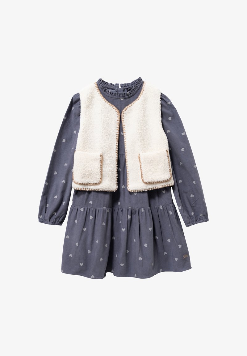 Granatowa sukienka w serca, z długimi rękawami i marszczoną spódnicą, nałożona na kremowy kamizelka z sztucznego futra z brązowymi wykończeniami i kieszeniami.