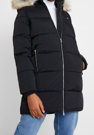 Personne portant une doudoune noire avec des poches zippées et une capuche bordée de fourrure, associée à un jean bleu et un haut blanc.