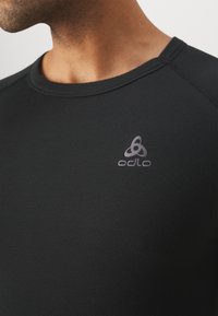 T-shirt de sport noir en tissu texturé, avec un col rond et un logo Odlo réfléchissant sur le haut à gauche.