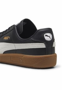 Sneaker neri in pelle con una striscia bianca, suola in gomma naturale e logo Puma dorato. Presenta un tacco arrotondato e una texture liscia.