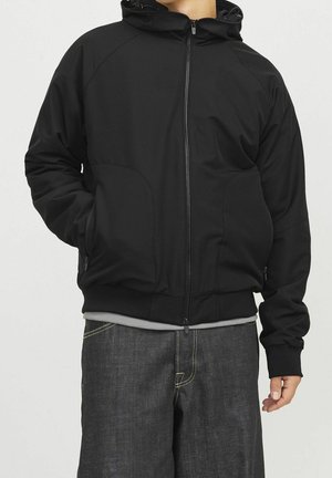 Man wearing a black zip-up hooded jacket with hands in pockets and dark denim jeans against a plain background.