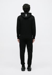 Vīrietis stāv, skatoties prom, valkājot melnu kapuci ar baltām svītrām kapucē, melnas sporta bikses un melnas kedas uz baltas fona.