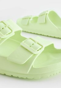 Sandales en plastique vert clair avec deux sangles réglables et des boucles rectangulaires. Semelle intérieure texturée et semelle plate et flexible.