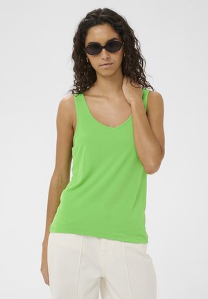 Donna con capelli ricci che indossa occhiali da sole neri, una canotta verde acceso e pantaloni bianchi, in piedi davanti a uno sfondo semplice.