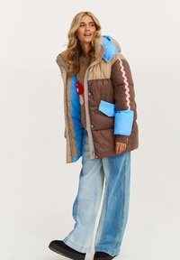 Žena v pufferovej bunde s farebnými blokmi v béžovej, hnedej a modrej farbe, skombinovanej s sveterom s motívom húb, voľnými džínsami a čiernymi topánkami.