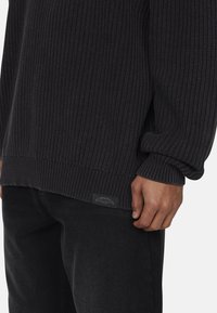 Maglione nero a maglia con texture a coste e vestibilità rilassata. Presenta un'etichetta sull'orlo inferiore e spalle scese. Abbinato a pantaloni neri.
