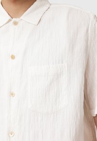Chemise blanche à manches longues en tissu texturé et froissé. Présente un col pointu et une poche poitrine unique avec des boutons beige.