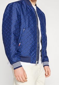 Blue bomber jacket with a textured geometric pattern, ribbed cuffs and waistband, zippered pocket, and a lightweight, smooth fabric finish.