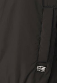 Čierna G-Star bunda s hladkým, matným povrchom. Disponuje nášivkou s logom na ľavom rúške a štruktúrovanými švami pozdĺž tela.