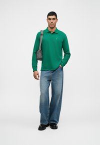 Zelená polokošela s dlhým rukávom, voľné svetlomodré džínsy, čierne topánky, drží sivý crossbody batoh, neutrálne pozadie.