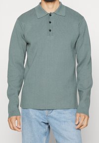 Polo à manches longues en tissu vert doux et texturé, avec un col et trois boutons noirs sur la patte de boutonnage.