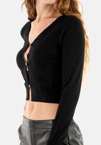Cardigan corto nero, in materiale lavorato a maglia, design a scollo a V, maniche lunghe, chiusura frontale con un singolo bottone metallico, abbinato a pantaloni in pelle grigia.